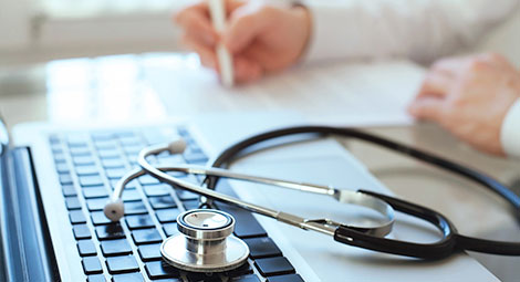 A stethoscope placed on a computer keyboard with a doctor writing in a chart in the background